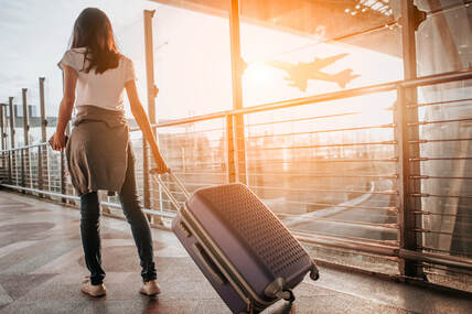 Picture of a woman pulling a rolling suitcase through the airport while an airplane is flying in the background